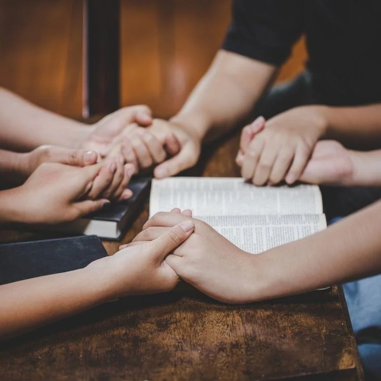People holding hands in prayer around an open Bible representing church fellowship.